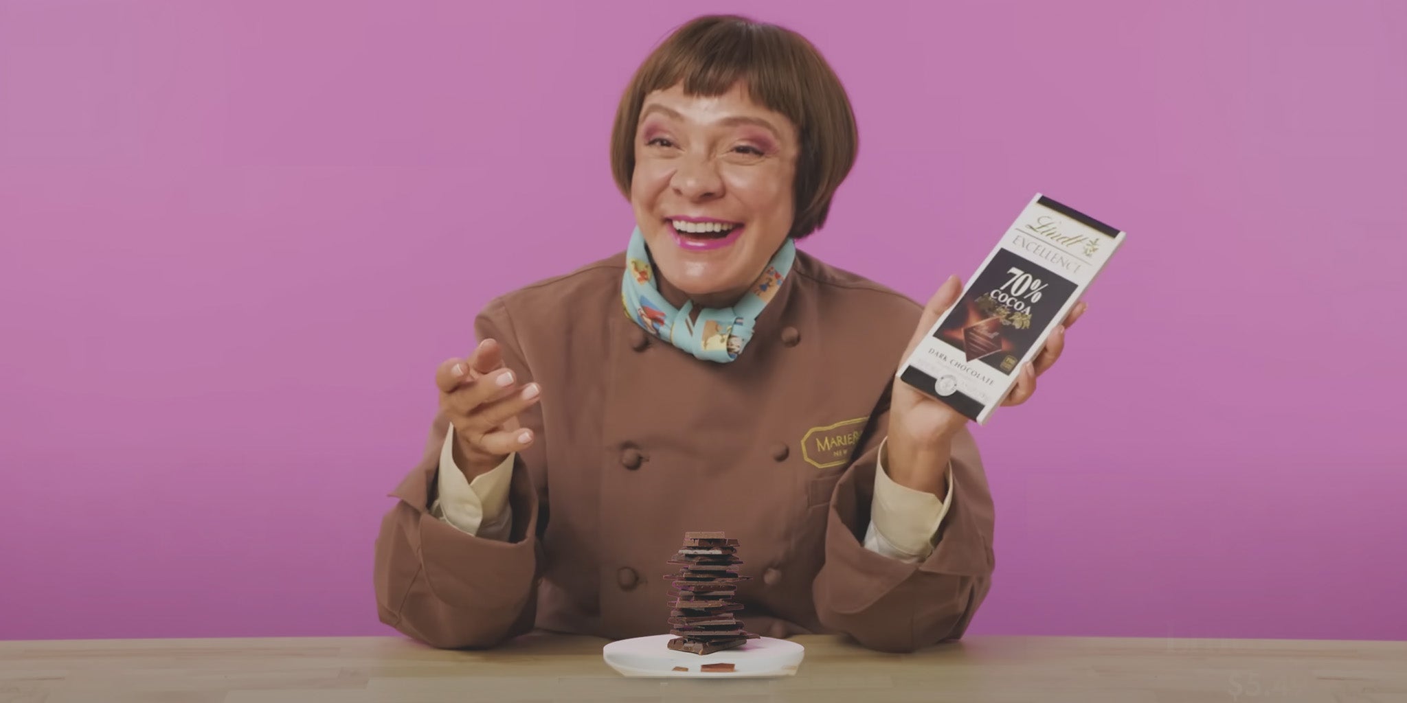 MarieBelle in a brown chef's coat holding a chocolate bar and a plate of chocolate pieces against a pink background