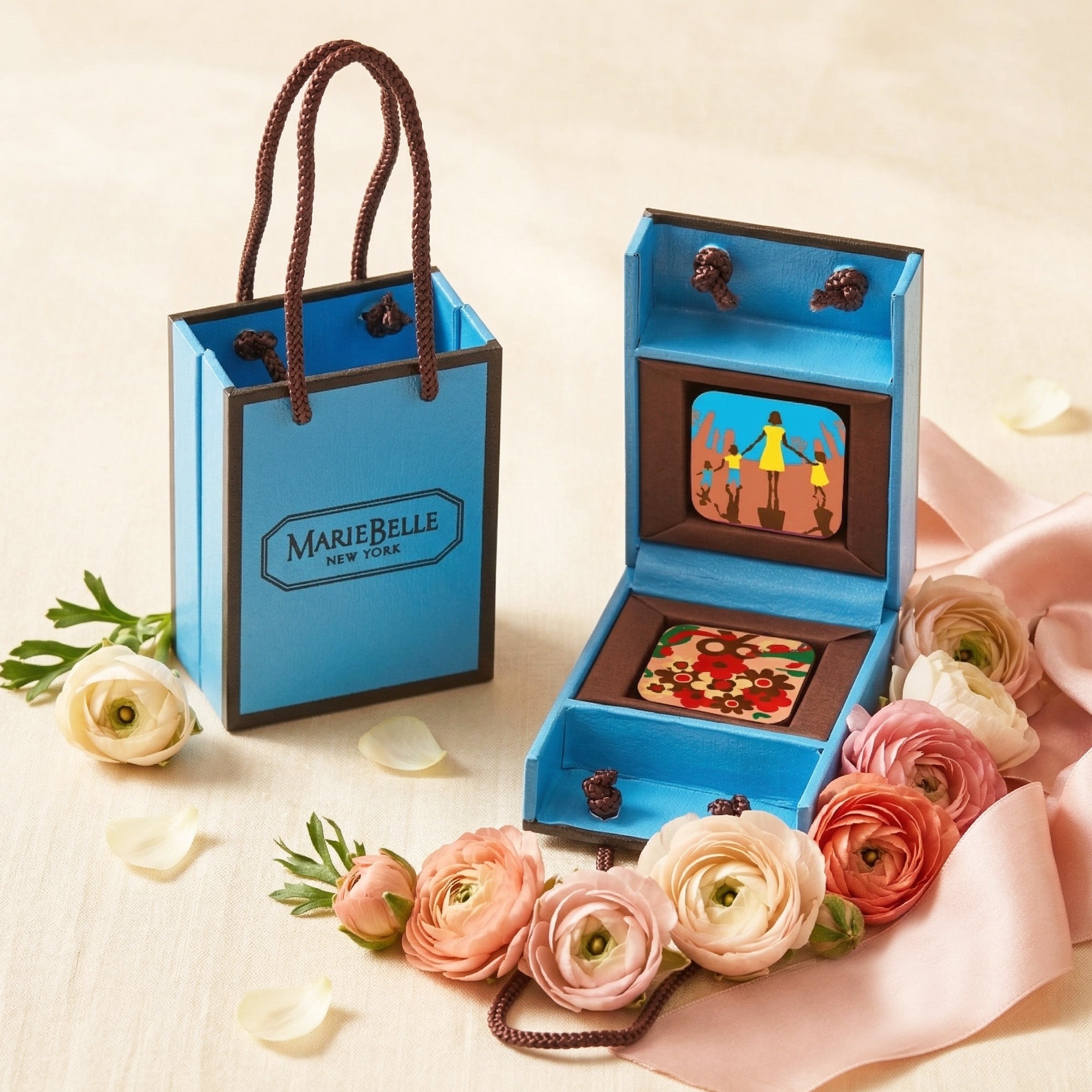 MarieBelle Blue gift bag with 'MarieBelle New York' branding next to a decorative box with a small figurine and flowers on a light background.