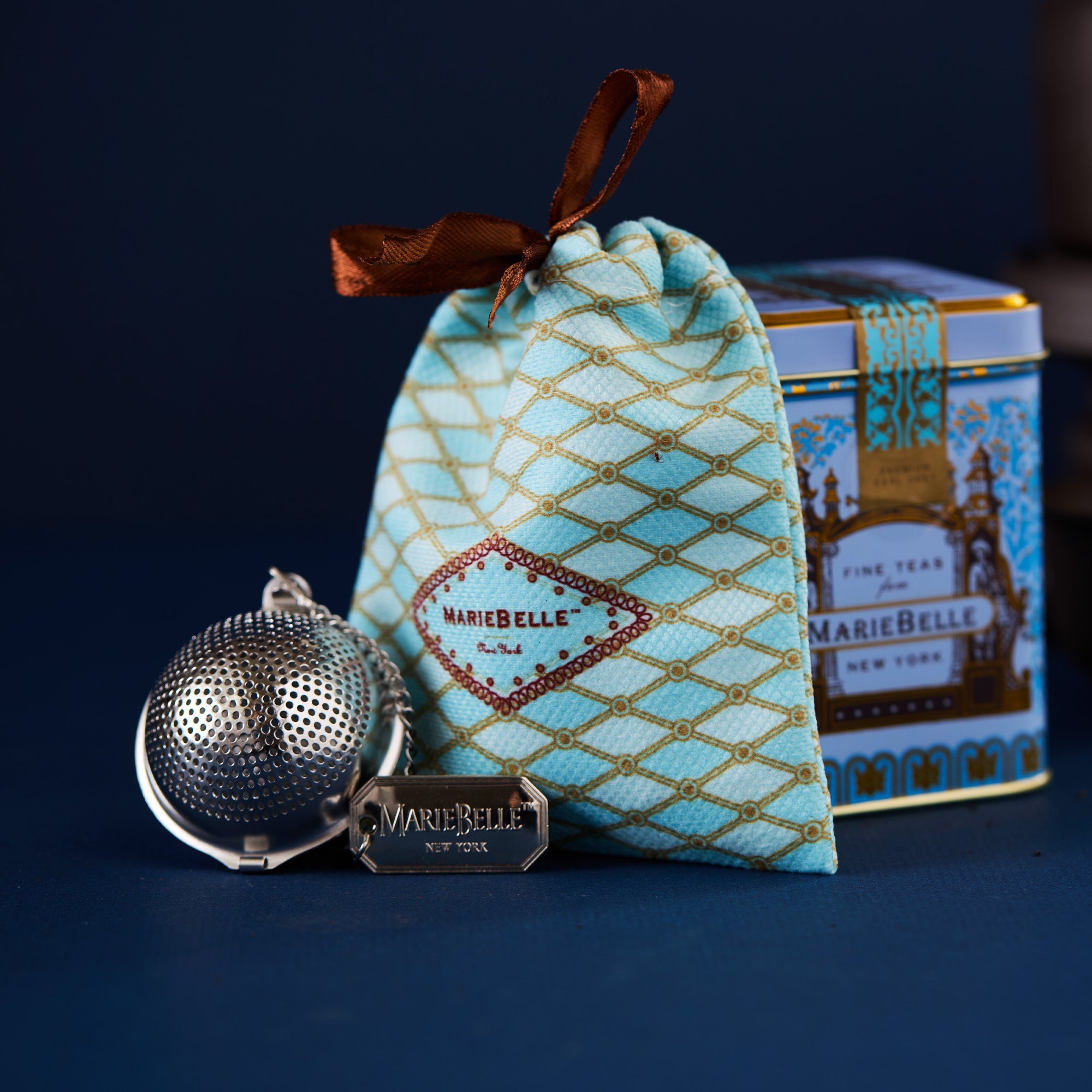Tea infuser, decorative pouch, and tea canister with 'MarieBelle' branding on a dark background